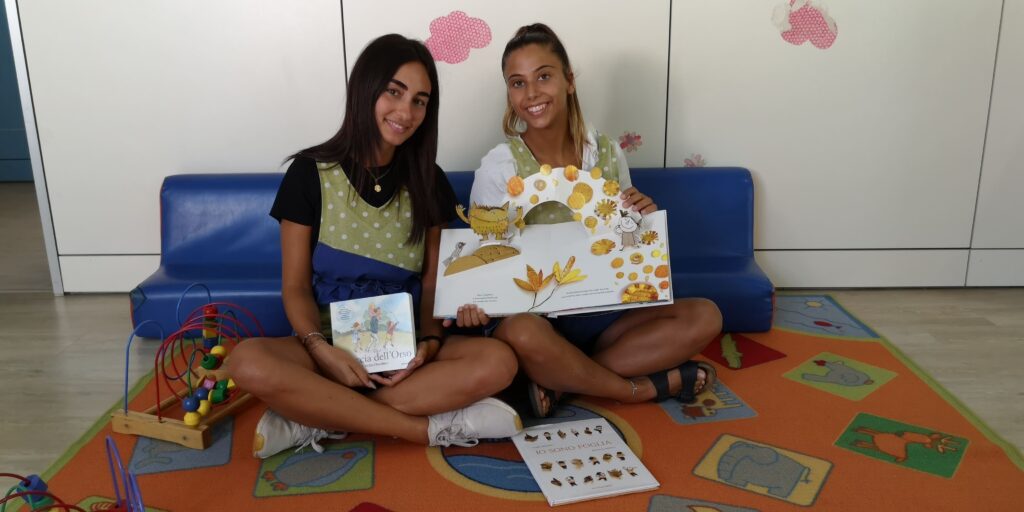 due ragazze sorridenti sedute per terra mostrano un libro educativo